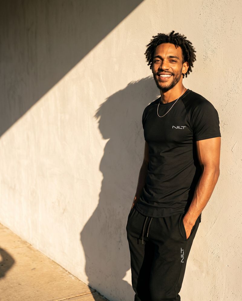 Homem encostado numa parede ao sol, de camiseta preta NILT e calça jogger